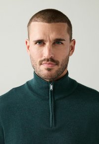 Mörkgrön zip-up-tröja med hög krage, tillverkad av ett mjukt, texturerat tyg. Har en silverfärgad dragkedja vid halsen.