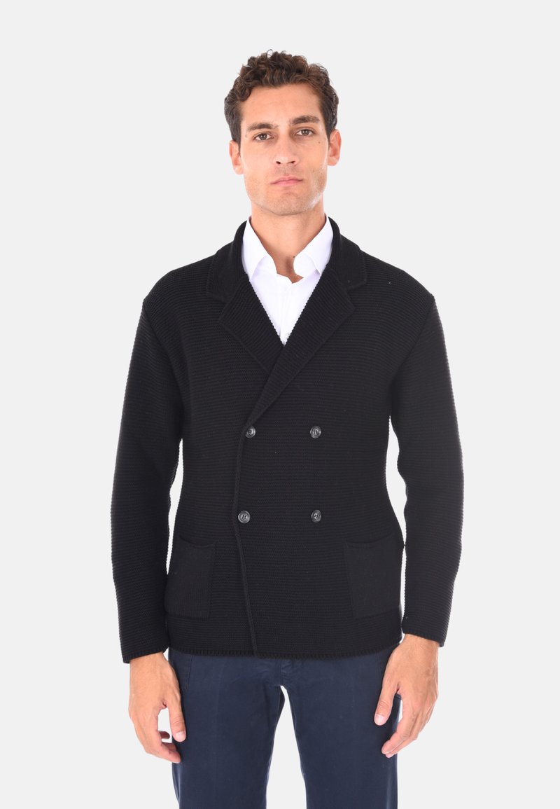 Cardigan nero doppiopetto, tessuto testurizzato, colletto a falda, due tasche frontali, bottoni evidenti, indossato su una camicia bianca.