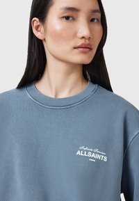 Modrá bavlněná mikina s kulatým výstřihem, zdobená bílým logem "ALLSAINTS" a textem. Materiál má hladkou strukturu s minimálními švy.