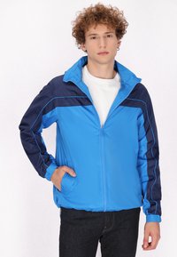Blauwe en marineblauwe windbreaker met een ritssluiting aan de voorkant, staande kraag, elastische cuffs en zijzakken; heeft contrasterende piping langs de schouders.