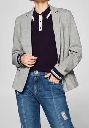 Personne portant un blazer gris sur un polo côtelé bleu marine avec une bordure blanche, associé à un jean bleu usé, une main dans la poche.