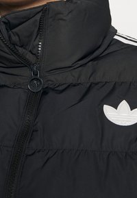 Black padded jacket with high collar, central zipper, white Adidas trefoil logo on chest, and three white stripes on shoulder.