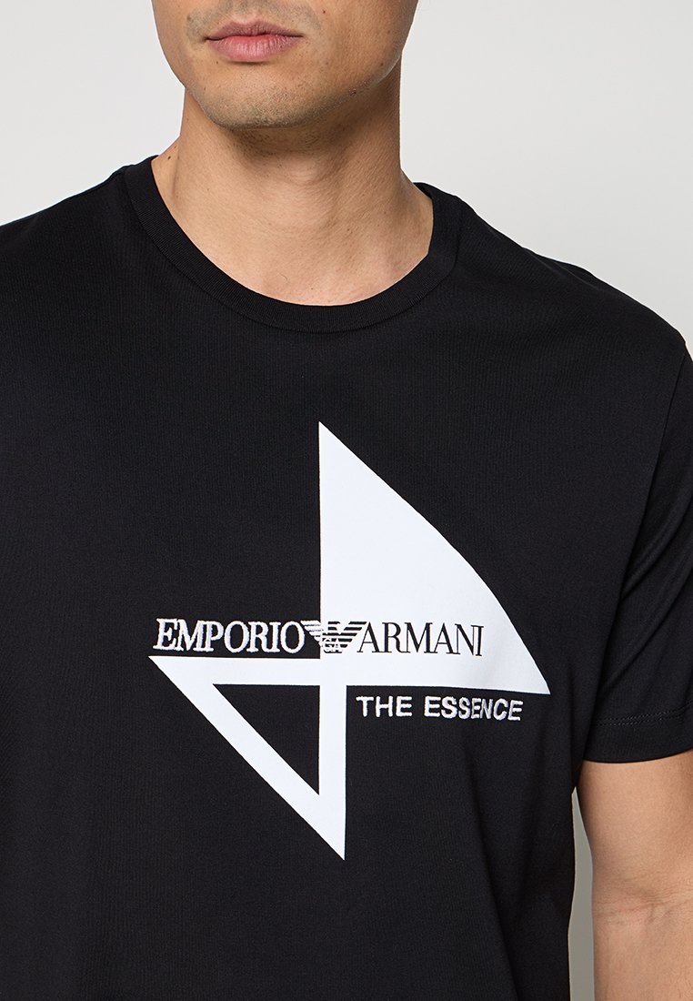 Hombre con camiseta negra con diseño geométrico blanco y texto "Emporio Armani The Essence" en el frente.