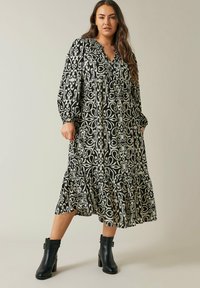 Schwarz-weiß gemustertes Midi-Kleid mit V-Ausschnitt, langen Ärmeln und gestuftem Saum. Kombiniert mit schwarzen Knöchelstiefeln, die eine strukturierte Oberfläche aufweisen.