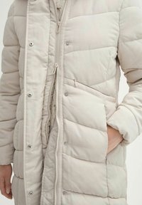 Veste matelassée beige clair avec une fermeture éclair à l'avant, des boutons-pression et une poche latérale. Présente une texture matelassée et un col montant pour plus de chaleur.