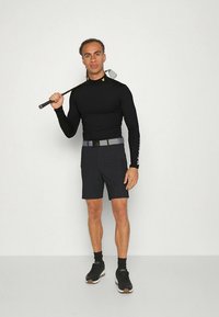 Črna majica z dolgimi rokavi in sive kratke hlače, obe tesno prilegajoči se. Na rokavu majice je logotip. Model drži golf palico in nosi črne športne čevlje.