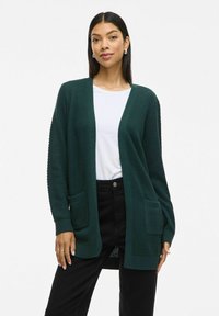 Temnozeleni pleteni kardigan z teksturiranim vzorcem, odprt spredaj, dolgi rokavi in sprednjimi žepi. Oblečen je čez belo majico in črne hlače.