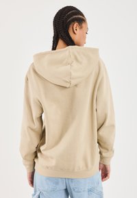 Person med fletninger iført en beige hættetrøje og lyseblå jeans, som vender bort fra en ensfarvet hvid baggrund.
