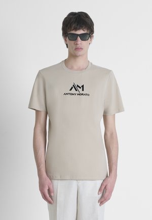 Beige katoenen t-shirt met een ronde hals, korte mouwen en zwart "ANTONY MORATO" logo in het midden. Het model draagt donkere zonnebrillen.