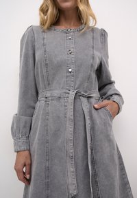 Gray denim dress with long sleeves, button-up front, and a belted waist. Features pleated shoulders and side pockets. Soft texture and structured shape.