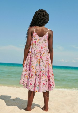 Fille aux cheveux tressés portant une robe rose à fleurs, debout pieds nus sur une plage de sable, face à la mer sous un ciel bleu clair.
