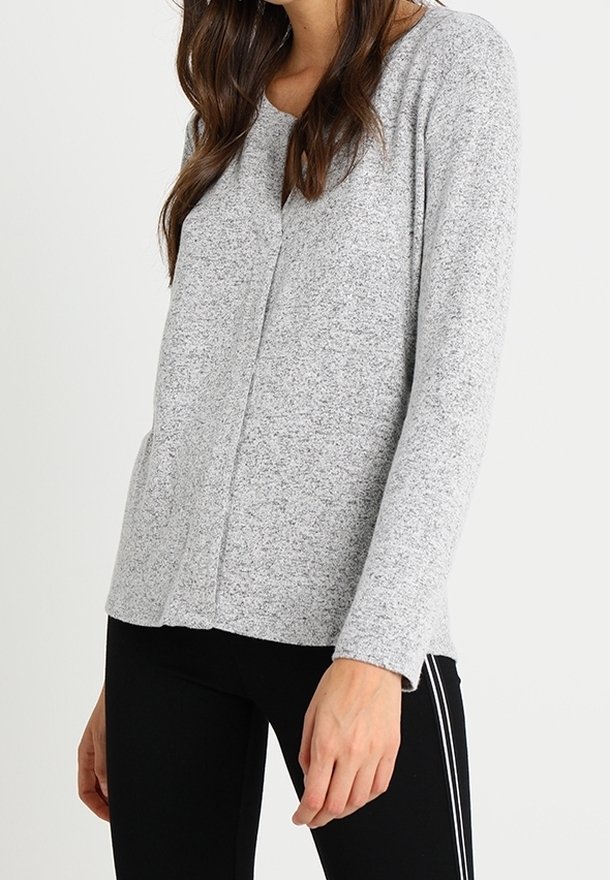 Femme portant un haut à manches longues gris clair et un legging noir avec des rayures blanches sur les côtés, se tenant devant un fond blanc uni.
