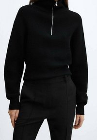 Pull en tricot noir avec un col haut et une demi-fermeture éclair. Design ajusté avec des manches longues et une texture côtelée. Assorti avec un pantalon noir.