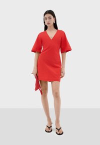 Vestido corto rojo con escote en V, mangas cortas acampanadas y un ajuste ceñido. Combinado con un clutch texturizado rojo y chanclas negras.