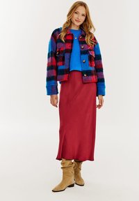 Wielokolorowa marynarka w kratę w odcieniach niebieskiego, czerwonego i czarnego, nałożona na niebieski sweter. Długa bordowa satynowa spódnica i beżowe, luźne botki dopełniają całość.