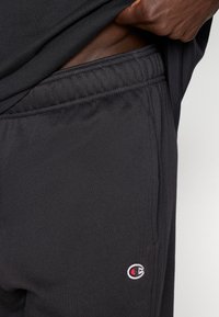 Μαύρα αθλητικά σορτς με ελαστική μέση, κατασκευασμένα από μαλακό ύφασμα, με ένα μικρό κόκκινο και λευκό λογότυπο στη αριστερή πλευρά.