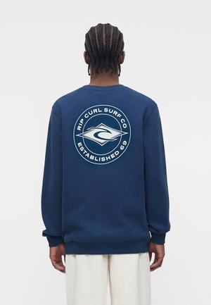 Osoba s vrkočmi oblečená v tmavomodrej mikine s bielym logom „Rip Curl Surf Co Established 69“ na chrbte, v kombinácii s krémovými nohavicami.