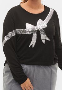 Schwarz langärmliges Oberteil mit einem silbernen Pailletten-Bow-Design auf der Brust, entspannter Schnitt und weicher Textur. Kombiniert mit grauen Hosen.