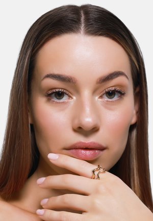 Goldfarbener Ring mit wellenförmigem Design, getragen an einer Hand mit hellrosa Nägeln, vor einem schlichten weißen Hintergrund. Glatte Haut mit natürlichem Make-up.