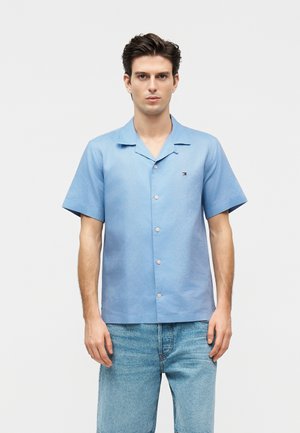 Junger Mann trägt hellblaues kurzärmeliges Hemd und blaue Jeans-Shorts, steht vor einem einfarbigen weißen Hintergrund.