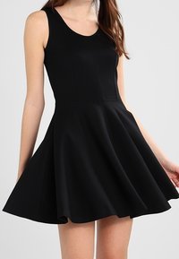Frau in einem ärmellosen schwarzen Skaterkleid mit tailliertem Oberteil und ausgestelltem Rock, vor einem schlichten Hintergrund stehend.