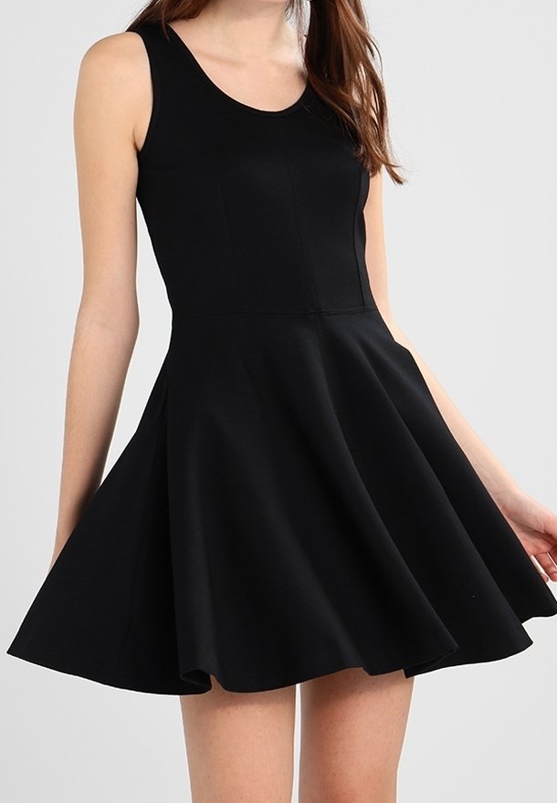 Frau in einem ärmellosen schwarzen Skaterkleid mit tailliertem Oberteil und ausgestelltem Rock, vor einem schlichten Hintergrund stehend.
