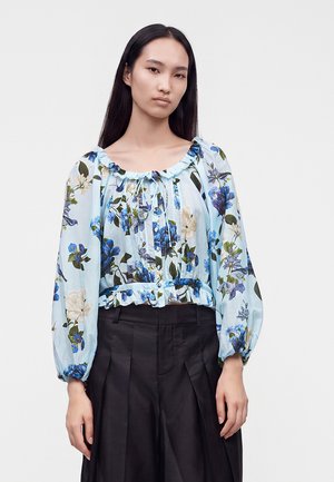 Femme portant un chemisier bleu clair à fleurs avec des manches longues bouffantes et un pantalon noir plissé taille haute sur un fond uni.