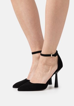 Jambes portant des escarpins noirs en daim à bout pointu avec brides autour de la cheville et boucles dorées sur fond blanc.