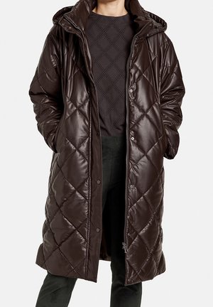 Lange donkergouden bruin doorgestikte pufferjas met capuchon, open gedragen over een zwart patroon top en donkere broek, handen in de zijzakken.