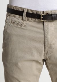 Beige Baumwollhosen mit strukturiertem Finish, ausgestattet mit einem dunkelbraunen Ledergürtel, seitlichen Taschen und einem klaren Schnitt entlang der Taille.