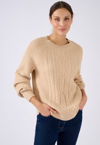Beige kabelgebreide trui met een wijde halslijn en geribde manchetten, voorzien van textuurpatronen en een relaxte pasvorm, gecombineerd met donkere jeans.