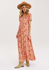 Ružová maxi šaty s oranžovými a žltými kvetmi, krátkymi rukávmi, priliehavým pásom a splývavou sukňou. Spárované s béžovými členkovými čižmami.