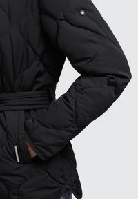 Giacca nera trapuntata con cuciture curve, tasca laterale con bottone a pressione e una cintura annodata in vita, indossata con una mano in tasca.