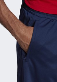 Granatowe spodenki sportowe o gładkiej fakturze, z elastycznym pasem i boczną kieszenią z zamkiem błyskawicznym. Czerwona koszulka widoczna na górze.