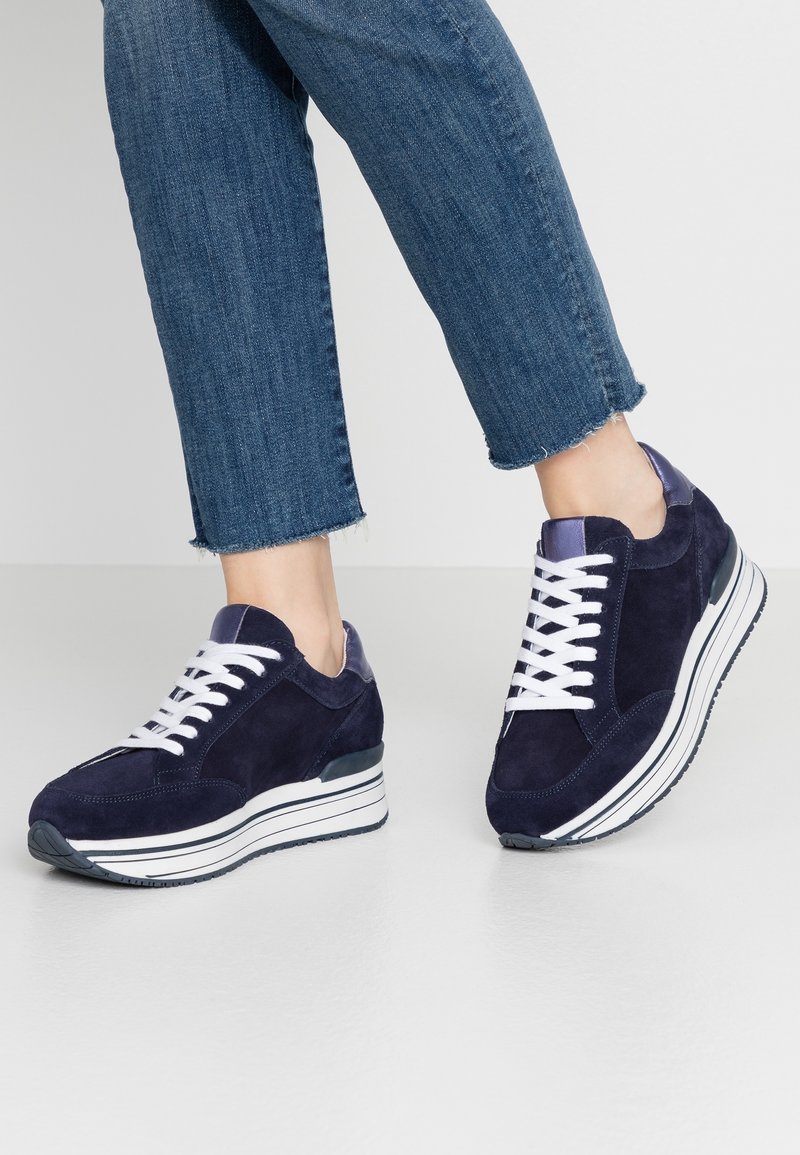 Pieds portant des baskets plateforme en daim bleu foncé avec des lacets blancs et des semelles rayées, associées à un jean bleu coupé.