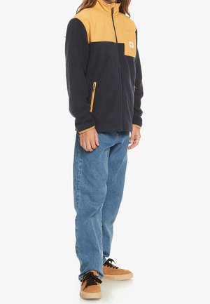 Personne portant une veste zippée noire et jaune, un jean bleu et des baskets beiges avec des lacets noirs, debout sur un fond blanc.