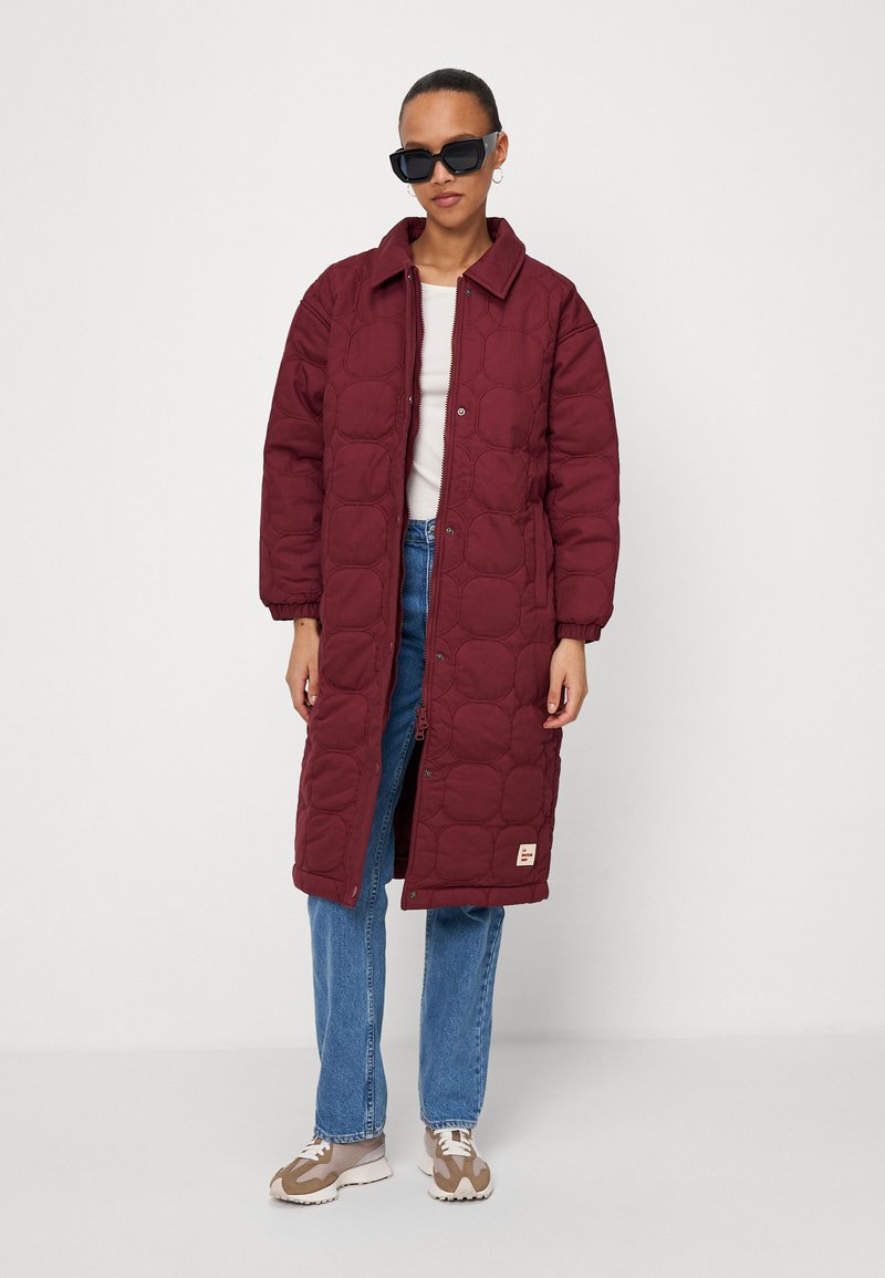 Manteau long matelassé bordeaux avec une fermeture à boutons, un col et des poignets élastiques ; porté sur un haut blanc et un jean bleu, associé à des baskets beiges.