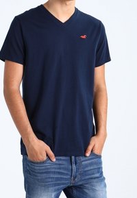 Man som bär en marinblå V-ringad T-shirt med en liten orange logotyp på bröstet och blå jeans, med händerna i fickorna.