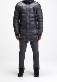 Man die een lange zwarte pufferjas draagt met ritszakken en donkergrijze broek, staand tegen een witte achtergrond.