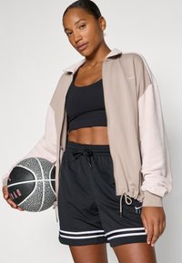 Lichtbeige zip-up jack met roze mouwen, zwarte crop top en zwarte shorts met witte strepen; houdt een zwart en grijs basketbal vast.