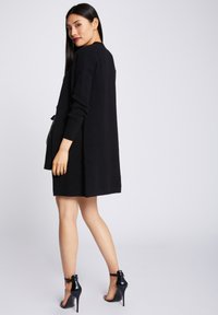 Gilet noir à manches longues en tissu doux, doté d'un design minimaliste et d'une coupe décontractée, associé à des chaussures à talons noirs.
