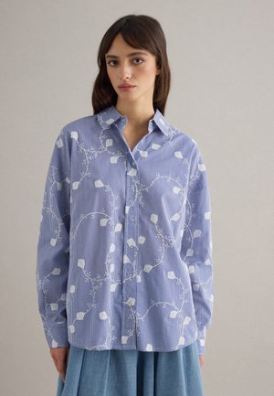 Donna che indossa una camicia a righe blu e bianche con ricami floreali bianchi, abbinata a una gonna azzurra, in piedi davanti a uno sfondo semplice.