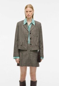 Grijze geruite blazer met een rechte snit, gecombineerd met een lichtblauw overhemd met kraag en een bijpassende rok. Accessoires omvatten een bruine clutch en kniehoge laarzen.