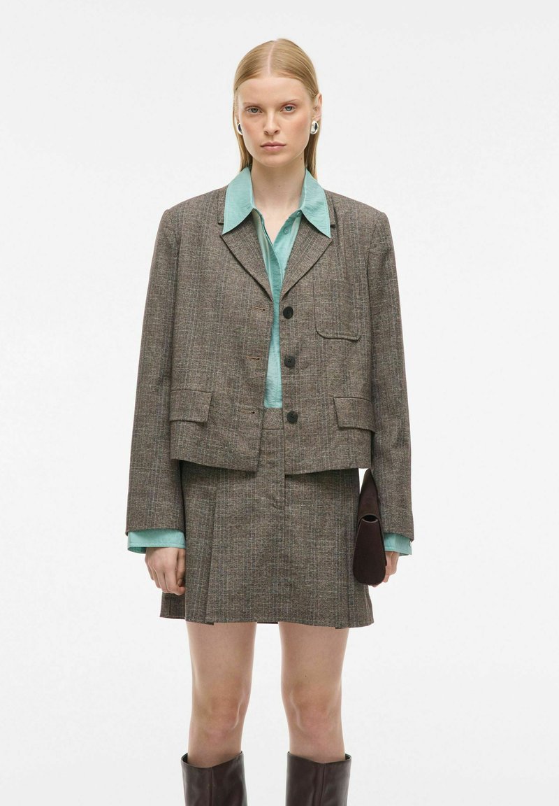 Grijze geruite blazer met een rechte snit, gecombineerd met een lichtblauw overhemd met kraag en een bijpassende rok. Accessoires omvatten een bruine clutch en kniehoge laarzen.