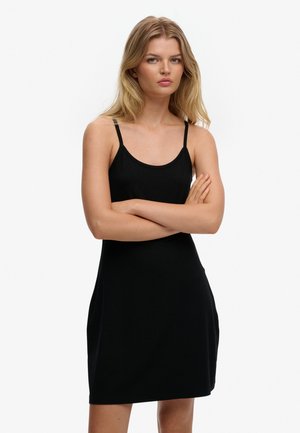 Jonge vrouw met blond haar draagt een mouwloze zwarte jurk met dunne bandjes, staand met gekruiste armen tegen een effen witte achtergrond.