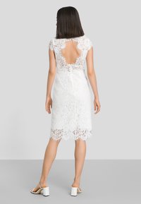 Robe en dentelle blanche avec des manches courtes, dos ouvert et ourlet festonné. La robe présente des motifs floraux complexes et une silhouette ajustée.