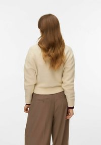 Bēšs adīts džemperis ar ribota apakšmalu, savienots ar brūniem platām biksēm ar aizmugurē esošām pogu detaļām. Vienkāršs, teksturēts dizains.