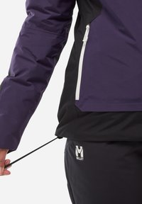 Veste imperméable violette et noire avec fermeture éclair et ourlet à cordon. Tissu texturé et logo Millet sur le pantalon noir.