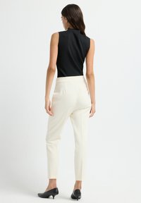 Vrouw die met de rug naar de kijker staat, gekleed in een mouwloze zwarte top, witte op maat gemaakte broek en zwarte lage hakken tegen een witte achtergrond.