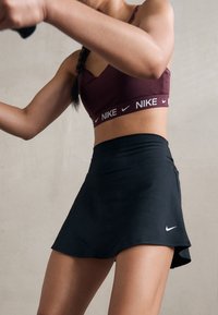 Falda atlética negra con un ligero vuelo y un logo de Nike. Combinada con un sujetador deportivo burdeos que presenta una banda de Nike. Textura de tela suave.